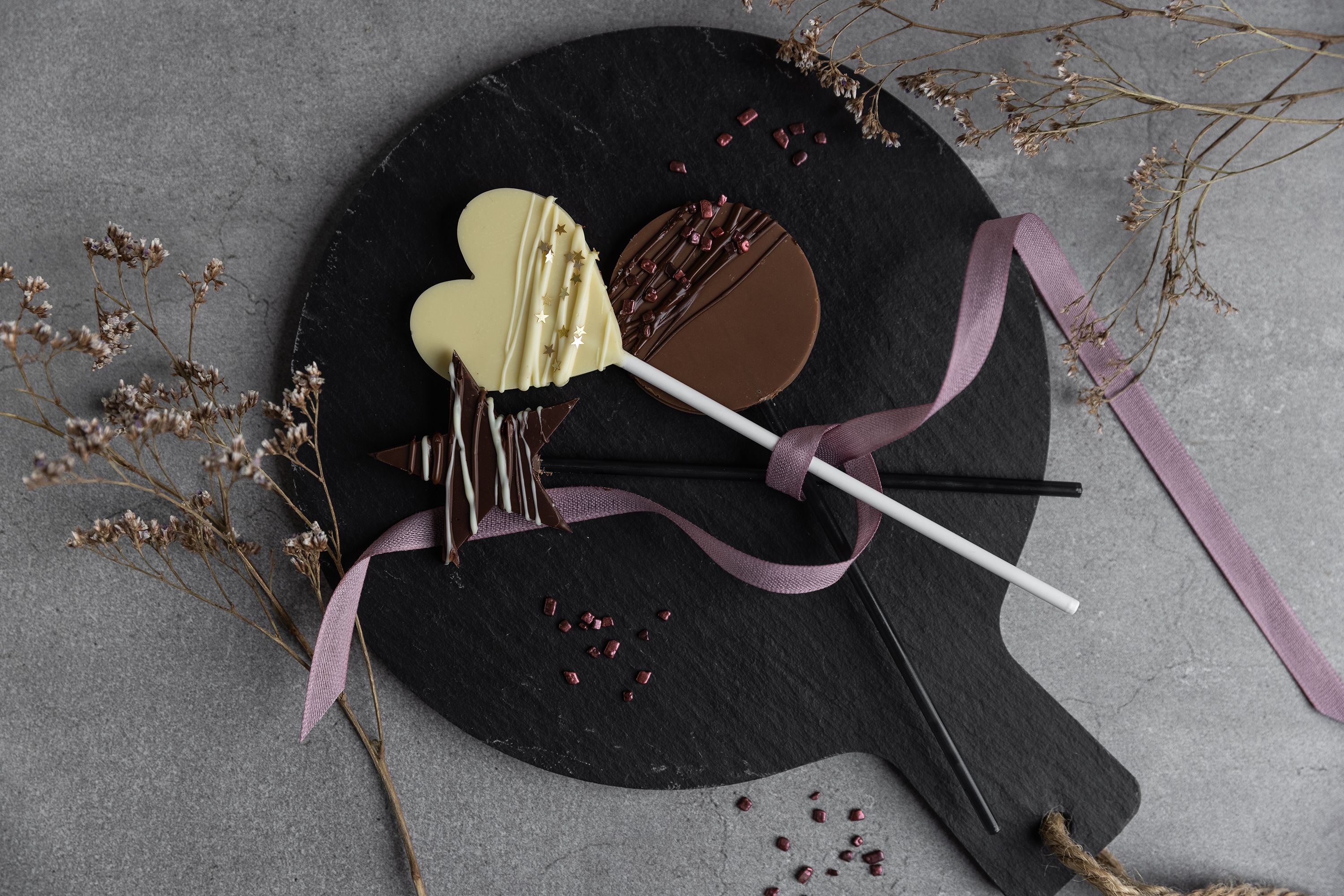 Schokoladenherz-Lutscher auf schwarzem Teller mit lila Schleife. 